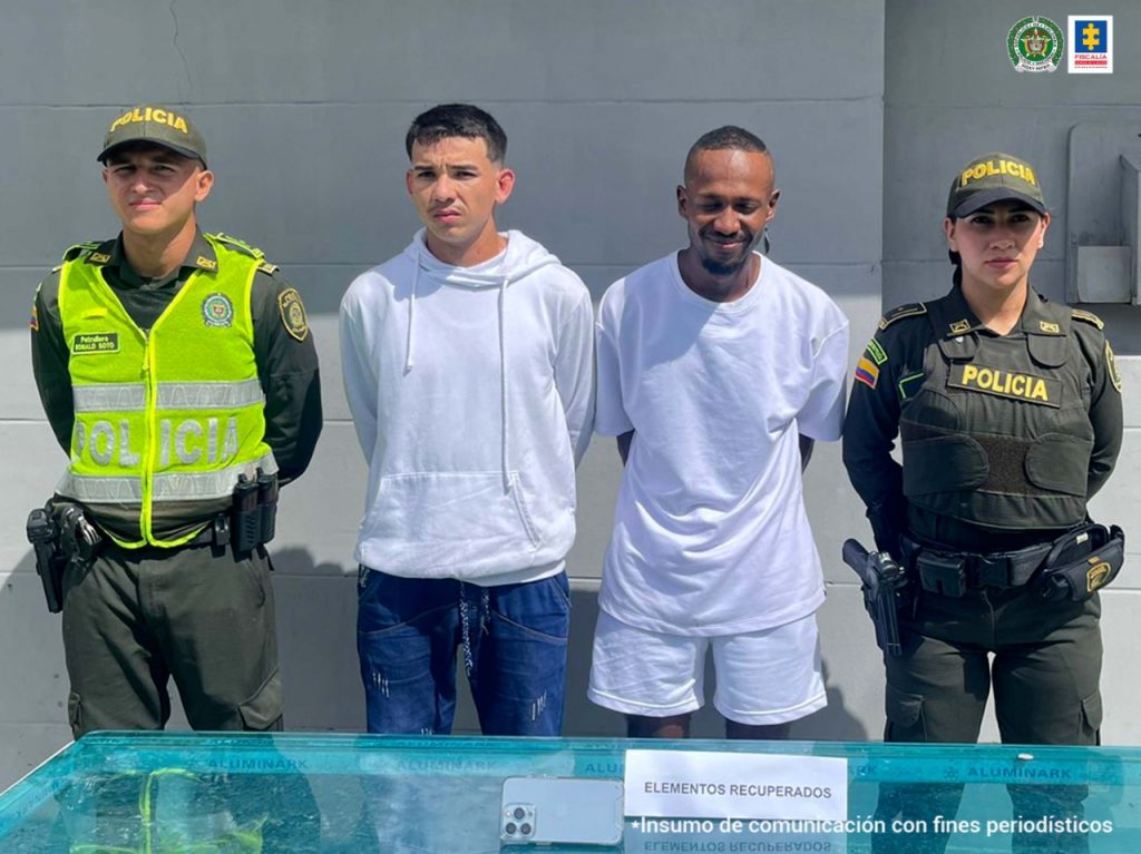 En la imagen se observa a dos hombres con camisetas blancas custodiado por dos agentes de la Policía Nacional.