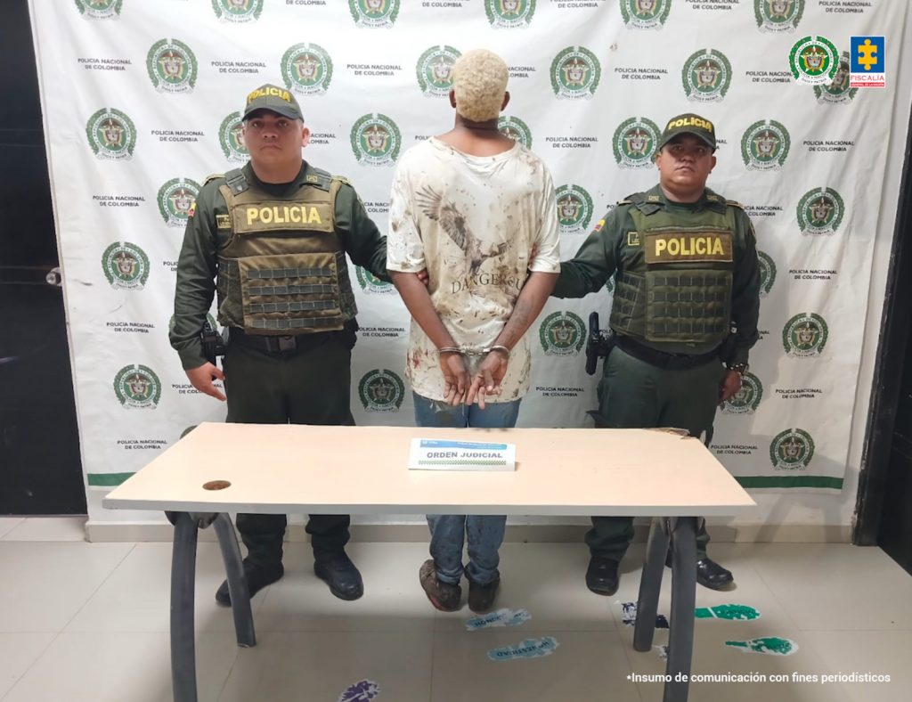 En la fotografía está un hombre de espalda con las manos esposadas, camiseta blanca, jean azul, cabellos rubios crespos, custodiado por dos uniformados de la Policía Nacional.