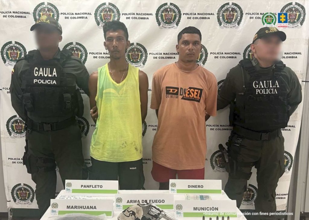 En la fotografía están dos hombres morenos de pie, con los brazos atrás esposados, uno tiene camisilla amarilla y el otro camiseta naranja. Se encuentran custodiado por dos uniformados de la Policía Nacional.