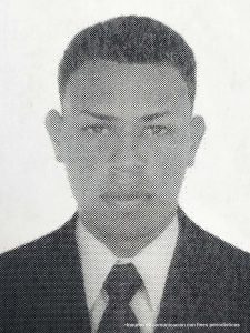 En la imagen a blanco y negro aparece el rostro de un hombre joven de ojos rasgados, nariz, boca y orejas grandes, cabellos crespos cortos, vestido con saco negro, camisa blanca y corbata negra.