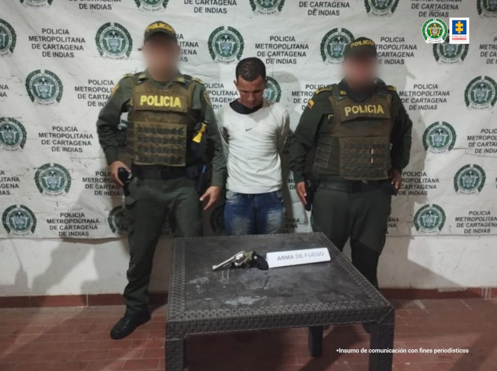 En la fotografía está un hombre de pie con la cabeza agachada, brazos atrás, vestido de buzo blanco y jean azul. Con él se encuentran dos agentes de la Policía Nacional custodiándolo. Delante de ellos hay una mesa con el arma incautada.