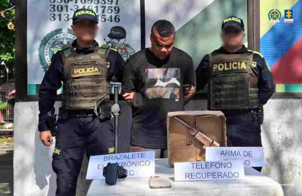 En la imagen está un hombre moreno de contextura mediana con la cabeza agachada de pelos cortos crespos, vestido con una camiseta negra en las afueras de un CAI de la Policía Nacional. A su lado están dos uniformados de la Policía custodiándolo.