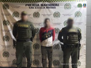 En la imagen se observa a un hombre de camiseta roja con rallas azul oscura y blanca con un jean negro custodiado por dos agentes de la Policía Nacional delante de un pendón de esa institución.