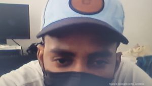 : En la foto aparece el rostro de un hombre trigueño, joven, de ojos grandes, con gorra azul, tapaboca negro y camiseta blanca, sentado en una silla asistiendo virtualmente a la audiencia en la que se le procesó.