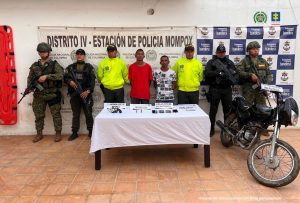 En la imagen están dos hombres de pie, ambos morenos, delgados, con los brazos atrás, acompañados por cinco uniformados de la Policía Nacional. Delante de ellos aparece una mesa con el arma, las municiones y el explosivo incautado.