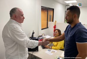 En la imagen se ven dos personas, de suéter blanco y azul, dándose la mano frente a una fiscal de suéter amarillo.
