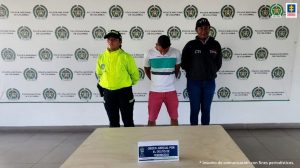  En la imagen se observa a un hombre de camiseta blanca con unas rayas, verde, beis y azul, bermuda roja, custodiado por un uniformado de la Policía Nacional y del CT, delante de un pendón de la Policia Nacional.
