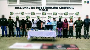 En la fotografía se aprecian de pie, en hilera y esposados entre sí a presuntos integrantes de ‘Los Escamers’. A los extremos hay servidores del CTI de la Fiscalía y uniformados de la Policía Nacional. Delante de ellos hay una mesa con mantel blanco y sobre ella se aprecian varios elementos como un computador portátil, una pantalla, un arma de fuego, celulares y sellos. Sobre el piso hay dos pendones con logos de una reconocida marca de motocicletas. Detrás de los hoy imputados hay un pendón institucional de la Policía.