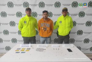 En la fotografía aparece de pie y con los brazos atrás uno de los presuntos integrantes de Los Manaos. Viste buso de manga larga color naranja con estampado; jean azul. A lado y lado de él hay uniformados de la Policía Nacional. Delante de ellos hay una mesa con mantel blanco y sobre ella hay tarjetas bancarias, munición y un celular. Asimismo, hay cuatro letreros que dicen: ‘Elementos incautados’, ‘Orden judicial’, ‘Munición’, ‘Celular incautado’. Detrás de ellos hay un pendón institucional de la Policía.