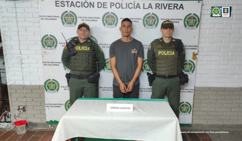 En la fotografía se aprecia a Diego Andrés Murillo Campo, de pie y con los brazos adelante y esposado. Viste camiseta de manga corta gris oscuro con un estampado pequeño al lado izquierdo del pecho; pantalón oscuro. A lado y lado de él hay uniformados de la Policía Nacional. Frente a ellos hay una mesa con mantel blanco y verde, sobre ella hay un letrero que dice: ‘Orden judicial’. Detrás de ellos hay un pendón institucional de la Policía.