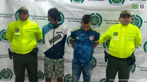 En la fotografía se aprecia a dos de los tres presuntos implicados en el hurto a la fundación de seguidores de un equipo de fútbol. Uno viste buzo de sudadera con franjas azul, blanca y negra y la marca ‘Puma’ en el torso; pantaloneta camuflada y pantuflas. La otra persona viste camisa de la Selección Colombia azul; jean con desgaste y zapatillas. A lado y lado de ellos hay uniformados de la Policía Nacional y detrás de ellos hay un pendón institucional de la Policía.