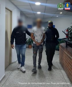 En la fotografía se aprecia en acción de caminata, en un pasillo, con los brazos adelante y esposado al judicializado. Viste camiseta de manga corta blanca con estampados negros; jean gris desgastado, pantuflas y medias negras. Al lado derecho de él hay un servidor del CTI de la Fiscalía; a la izquierda de él un uniformado de la Policía Nacional.