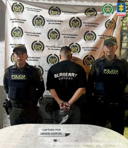 En la fotografía se aprecia de pie, con los brazos atrás, esposado y de espaldas a la cámara  a Juan David Sarria Zúñiga. Viste camiseta negra de manga corta y  pantalón negro. A sus extremos hay uniformados de la Policía Nacional. Sobre una superficie blanca hay un  letrero que dice: ‘Captura por orden judicial’. En la parte posterior, hay un pendón institucional de la Policía. 