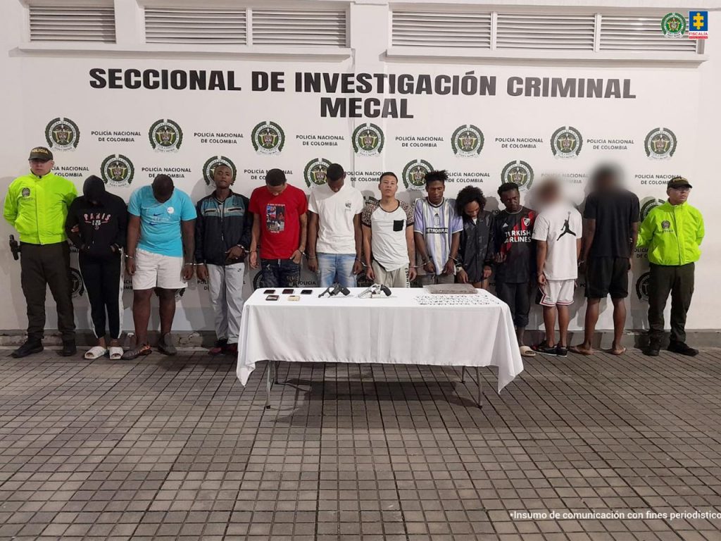 En la fotografía aparecen en hilera y esposados entre sí los presuntos integrantes de ‘La Ocho’. A los extremos hay uniformados de la Policía Nacional. Delante de ellos hay una mesa con mantel blanco y sobre ella se aprecian armas de fuego, cigarrillos, una bolsa de plástico con contenido vegetal y celulares. Detrás de ellos hay un pendón institucional de la Policía.