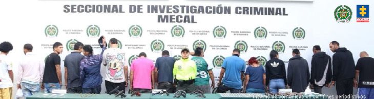 En la foto se aprecia en hilera y de espaldas a la cámara a los presuntos integrantes de ‘Las Veraneras. En la parte central de la fotografía hay una uniformidad de la Policía Nacional. Delante de ellos se alcanza a visualizar una superficie verde y sobre ella varios elementos como armas de fuego, munición, entre otros. En la parte posterior, hay un pendón institucional de la Policía.