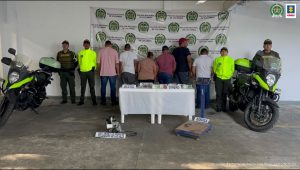 En la foto aparecen de y de espaldas a la cámara los presuntos integrantes de ‘Los Contaminadores’. A lado y lado de ellos hay uniformados de la Policía Nacional. Delante de ellos hay una mesa con mantel blanco y sobre ellas varios elementos y letreros que dicen: ‘Arma de fuego’, ‘Celulares’, ‘Dinero’. Asimismo, en el piso se observa una báscula y un aparato junto a un letrero que dice ‘Selladora de costales’. En la parte posterior hay un pendón institucional de la Policía.