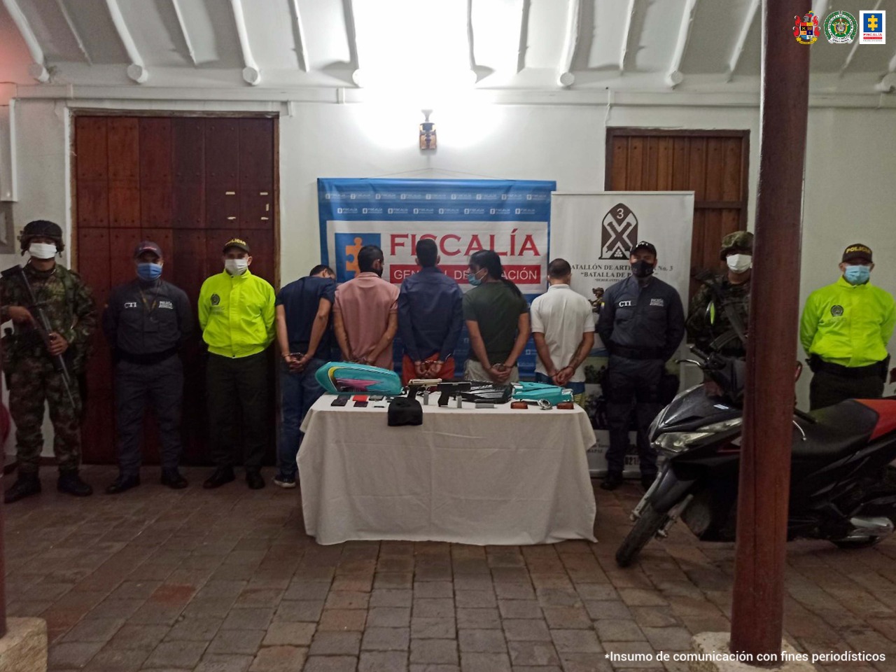 Aparecen los presuntos integrantes del grupo delictivo ‘Los Lobos’, de espalda. A lado y lado hay servidores del CTI de la Fiscalía, uniformados de la Policía Nacional y del Ejército. Delante de ellos hay una mesa con mantel blanco y sobre ella varios elementos como armas de fuego y otros. Detrás de ellos hay dos pendones institucionales de la Fiscalía y del Ejército.