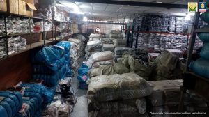 En la fotografía se aprecia una bodega con estanterías y en ellas gran cantidad de paquetes en plástico y cajas. Asimismo, en el piso hay paquetes, uno sobre otros, envueltos en plásticos transparentes, de color azul y amarillo. 