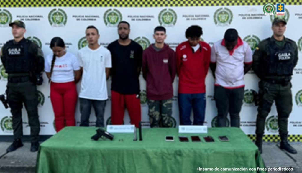 En la fotografía se aprecia en hilera y con los brazos atrás, a los seis judicializados, entre los que hay una mujer. A sus extremos hay uniformados del Gaula de la Policía Nacional. Delante de ellos hay una mesa con mantel verde oliva y sobre ella hay un arma de fuego, un proveedor y varios teléfonos celulares. Detrás de ellos hay un pendón institucional de la Policía.