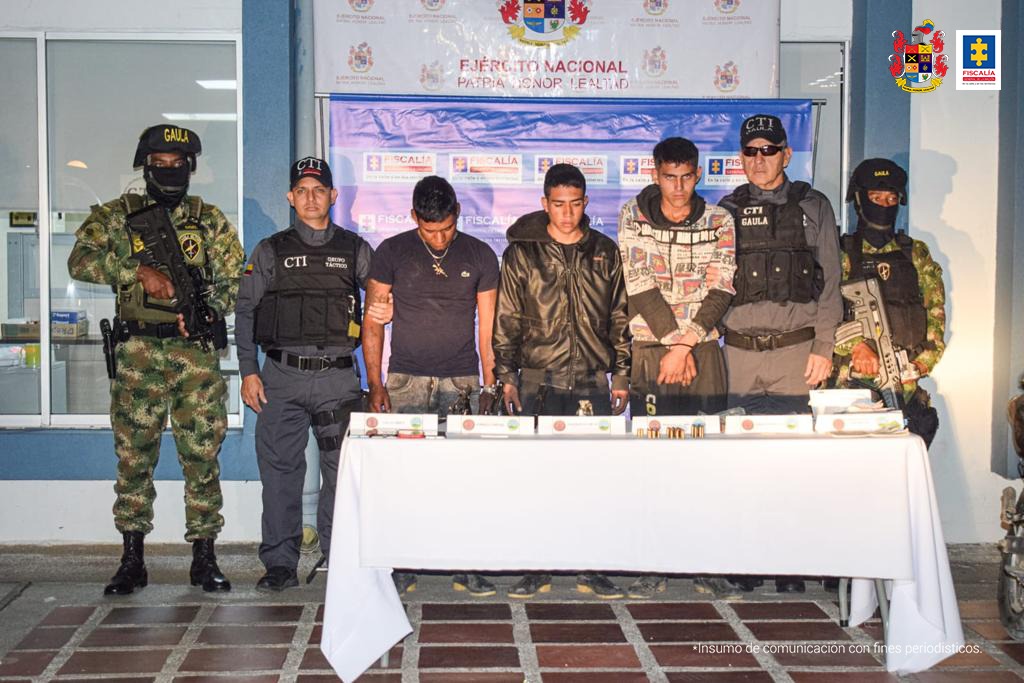 En la fotografía se aprecian los tres presuntos integrantes del Gaor Ismael Ruiz de las Farc EP. A lado y lado de ellos hay servidores del CTI de la Fiscalía y del Ejército Nacional. Delante de ellos hay una mesa con mantel blanco y sobre ellas unos elementos. Detrás de ellos hay dos pendones institucionales de la Fiscalía y del Ejército.