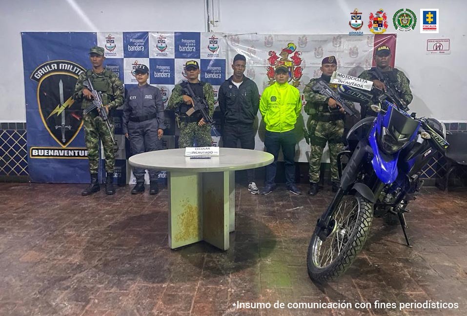 En la foto aparece Ronal Alejandro Aldana Cáceres, alias ‘Morocho’, con los brazos atrás, vestido con chaqueta y pantalón oscuros. A lado y lado de él hay uniformados del CTI de la Fiscalía, Policía, Armada y Ejército Nacional. Delante de ellos hay una mesa redonda y sobre ella un teléfono celular y un letrero que dice: ‘celular incautado’. Al lado izquierdo de la mesa hay una motocicleta. Detrás de ellos hay dos pendones institucionales del Gaula Militar de Buenaventura y del Ejército Nacional.