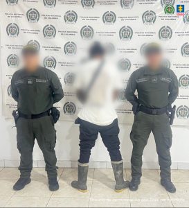 El capturado se encuentra de espaldas y custodiado por dos uniformados de la Policía Nacional. Detrás de la foto hay un backing de la Policía Nacional y el capturado tiene un buzo blanco, jeans y botas.
