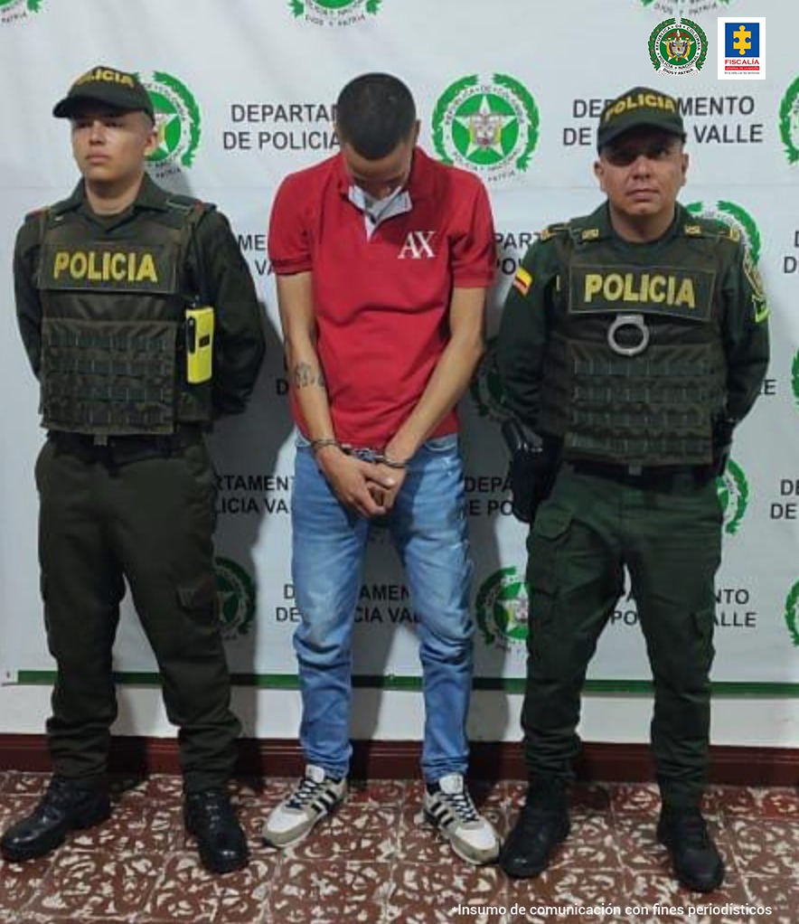 En la foto aparece de pie, con los brazos adelante y esposado, la cabeza agachada. Está vestido con camiseta roja, jean azul y zapatillas. A lado y lado de él hay dos uniformados de la Policía Nacional. Detrás de ellos hay un pendón institucional de la Policía Nacional. 