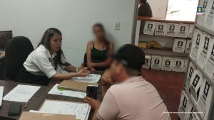 En la imagen se ve una funcionaria de la entidad de blusa blanca, atendiendo a dos usuarios, uno de ellos tiene una gorra negra. Ellos se encuentran dentro de una de las oficinas de la entidad.