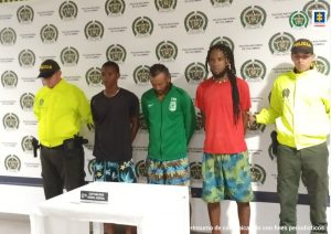 En la imagen aparecen tres hombres morenos, uno vestido de camiseta negra y bermuda estampada, el otro con camiseta verde y bermuda estampada y el otro con camiseta roja y bermuda estampada, custodiados por dos agentes de la Policía Nacional.