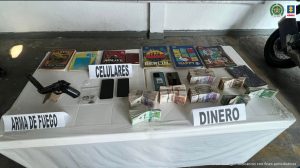 En la foto aparece una mesa con mantel blanco y sobre ella se observa, un arma de fuego, cuatro teléfonos celulares, dinero en efectivo y varios cuadernos.