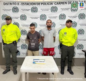 En la imagen están dos hombres de pie, uno está vestido con camiseta café y bermuda beis y el otro con camiseta gris y bermuda roja. Se encuentran acompañados de dos agentes de la Policía Nacional y detrás de ellos hay un pendón que dice Estación de Policía Nacional El Carmen de Bolívar.