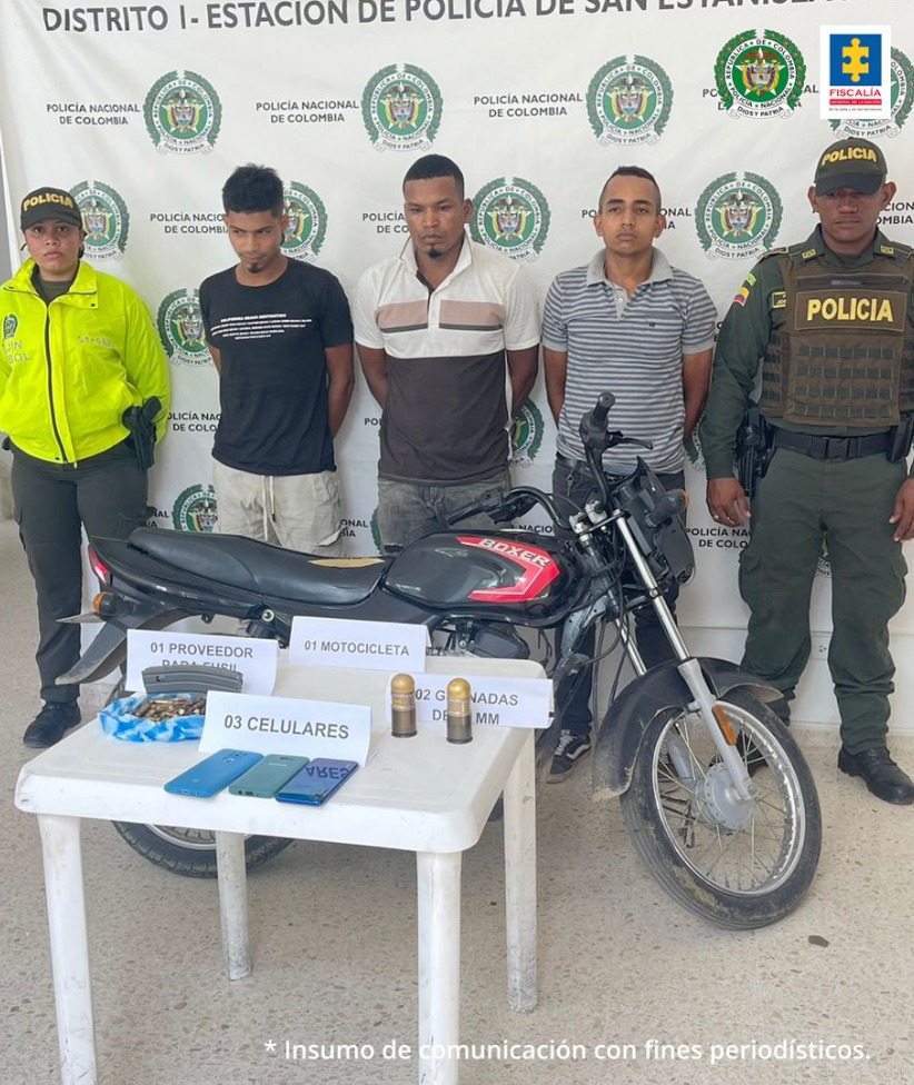 En la imagen hay tres hombres de pies, todos con los brazos atrás. Uno esta vestido con camiseta negra y pantalón beis, el otro está con una camiseta marrón con blanco y jean gris y el otro con camiseta gris de rallas y jean azul, custodiados por dos uniformados de la Policía Nacional. Delante de ellos hay una mesa blanca que tiene encima celulares, municiones y proveedores.