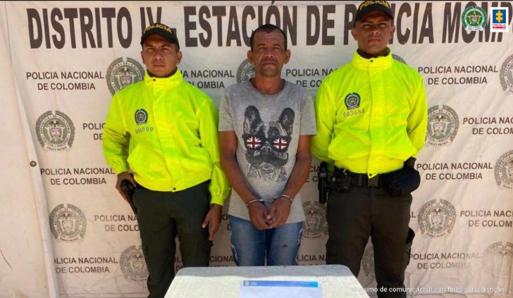 En la fotografía está un hombre moreno con las manos esposadas, vestido de camiseta gris, jean azul, acompañado de dos uniformados de la Policía Nacional. Detrás de ellos hay un pendón que dice Distrito IV, Estación de Policía Mompox.