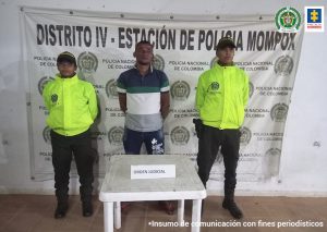 La fotografía muestra a un hombre moreno con camiseta de rallas azul con verde y blancas y jean azul oscuro, custodiado por dos agentes de la Policía Nacional delante de un pendón de esa institución.