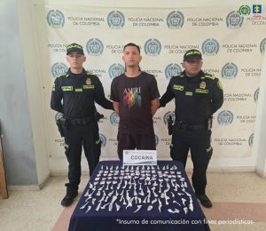 En la fotografía esta un hombre trigueño, joven, de pie, de frente, con los brazos atrás esposados, cabellos negros semi largo, está vestido con una camiseta y pantalón negro. Se encuentra acompañado de dos policías y delante de ellos hay una mesa verde con la droga incautada. 
