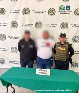 En la imagen aparece un hombre de contextura gruesa, de pie, de frente con los brazos atrás esposados, su rostro se encuentra difuminado y está vestido con camiseta blanca, jean azul. Está acompañado de un uniformado del CTI y otro de la Policía Nacional. 