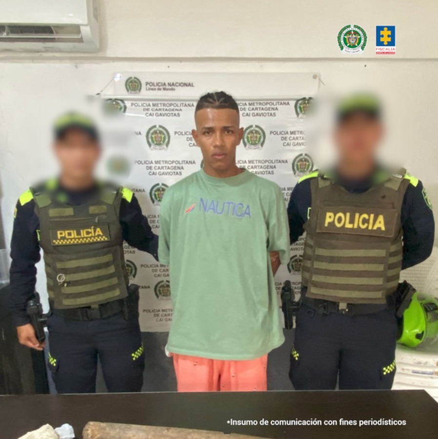 En la fotografía aparece un hombre joven moreno, de pie, delgado con los brazos atrás esposados, vestido con una camiseta verde y una bermuda rosada. Se encuentra custodiado por dos uniformados de la Policía Nacional.