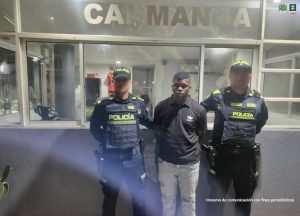 En la imagen esta un hombre de pie, de frente, moreno, cabellos crespos negros, vestido con un buzo negro y un jean azul. A su lado se encuentran dos policías y están en las afueras de un CAI. 