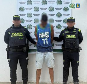 En la imagen está un hombre delgado de pie, de frente, de alta estatura, con la cabeza agachada, el rostro difuso, vestido con una camisilla azul y una bermuda azul clara de jean. Se encuentra custodiado por dos policías. 