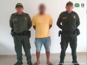 En la imagen está un hombre de pie, con el rostro difuso, los brazos atrás, vestido de camiseta amarilla y bermuda de jean azul con sandalias negras. Se encuentra acompañado de dos agentes de la Policía Nacional.