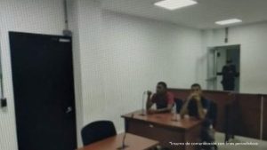 Imagen de audiencia. En la foto están dos hombres morenos sentados en un escritorio de madera, uno con camiseta roja, otro con camiseta negras, con micrófonos al interior de la sala donde se realizó la audiencia en la que se les judicializó.