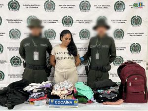 En la imagen hay una mujer de pie, con los brazos atrás, trigueña, de pelo largo negro, vestida con camiseta y sudadera beige y está acompañada de dos uniformados de la Policía Nacional. Delante de ellos hay una mesa con la ropa que se le incautó.