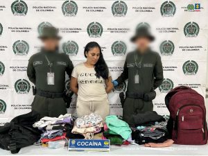     En la imagen vemos a una mujer de pie con los brazos a la espalda, de pelo negro, pelo largo y negro, vestida con camiseta y sudadera de color beige y acompañada de dos miembros uniformados de la Policía Nacional.  Frente a ellos hay una mesa con la ropa incautada.