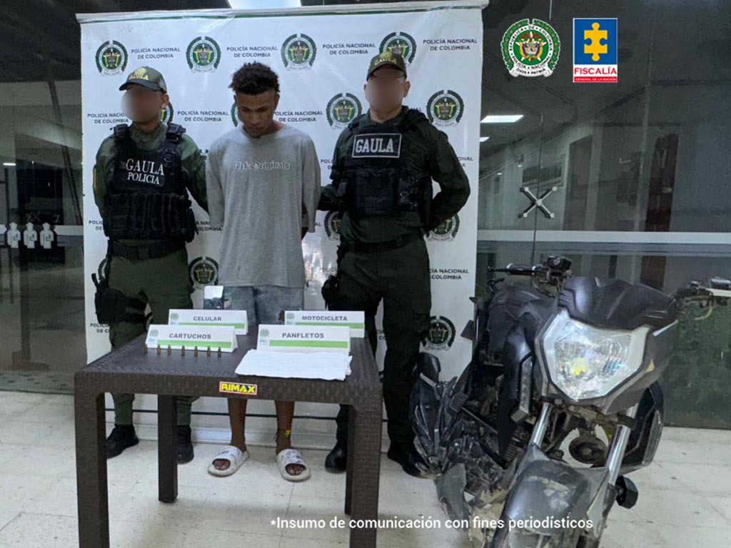 En la imagen esta un hombre alto, delgado, moreno, con los brazos atrás, vestido de camiseta gris con bermuda de jean azul y sandalias blancas, custodiado por dos uniformados de la Policía Nacional. 