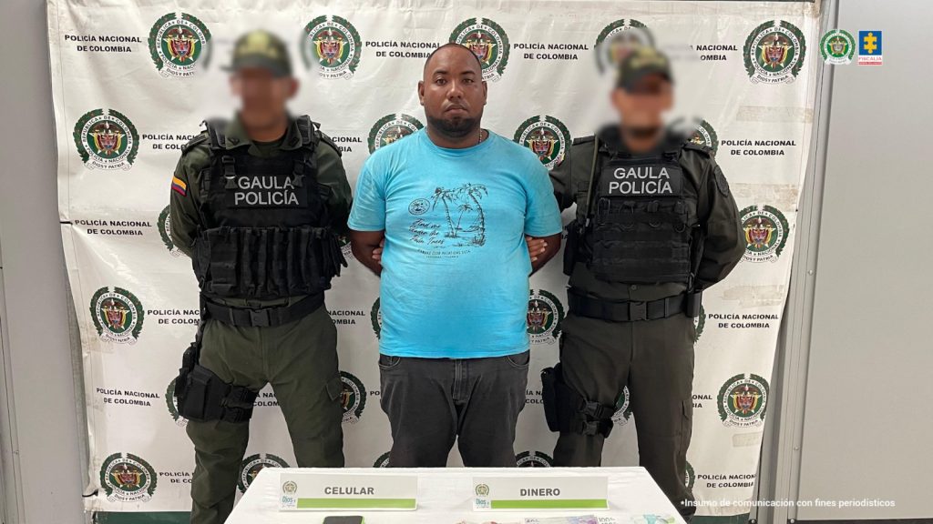 En la imagen está un hombre moreno, de pie, con los brazos atrás esposados, de contextura gruesa, abdomen pronunciado, barba negra, semi calvo, vestido con camiseta azul y jean negro. A su lado están dos uniformados de la Policía Nacional.