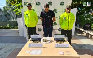 En la fotografía se observa a un hombre vestido de camisa y pantalón negro, custodiado por dos agentes de la Policía Nacional, delante de un pendón de esa institución. También se divisa una mesa blanca con la droga que le fue decomisada.