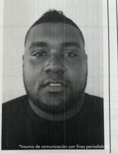 En la imagen a blanco y negro está el rostro de un hombre moreno, de contextura gruesa, con barba, cabellos cortos negros, cejas pobladas, ojos pequeños, boca mediana que se encuentra vestido con una camiseta negra.