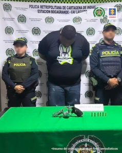 En la imagen hay un hombre moreno de contextura gruesa con la cabeza agachada y los brazos atrás. Se encuentra vestido con un buzo azul oscuro manga larga y un jean azul, custodiado por dos uniformados de la Policía Nacional. Delante de ellos hay una mesa con mantel verde donde hay un arma de fuego.
