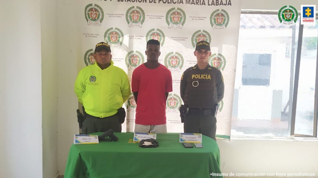 En la fotografía se observa a un hombre de tez morena con camiseta roja, custodiado por dos agentes de la Policía Nacional delante de un pendón de esta institución y con una mesa que muestra el arma y los celulares incautados.