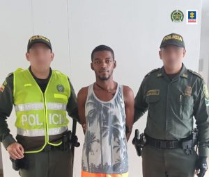En la imagen hay un hombre moreno vestido con camisilla estampada gris, se encuentra con los brazos atrás y está acompañado de dos agentes de la Policía Nacional.